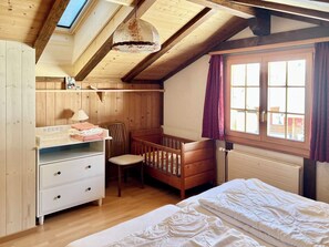 Une chambre avec un lit superposé, un berceau, un bureau avec des tiroirs, une chaise et une fenêtre avec des rideaux.