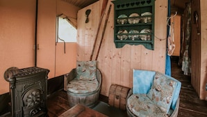 A rustic room with wooden walls, a window, a chair with a patterned cushion, a wooden barrel, and a shelf with glassware. - Kampari - Luxe Safaritent - Titanic by Interhome (Delfstrahuizen)