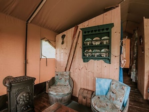 A rustic room with wooden walls, a window, a chair with a patterned cushion, a wooden barrel, and a shelf with glassware. - Kampari - Luxe Safaritent - Titanic by Interhome (Delfstrahuizen)