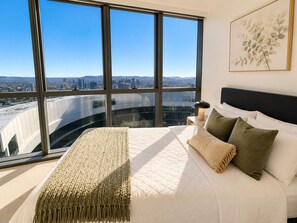 Une chambre avec une grande fenêtre, un lit recouvert de draps blancs et une vue sur la ville.