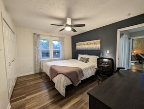 Un dormitorio con cama, ventilador de techo, ventana con cortinas, un cuadro en la pared y un escritorio con silla.