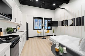 A modern kitchen with white cabinets, a stove, and a sink. There is a dining area with two chairs and a table. A bed is visible in the background. - Make Memories Downtown | Walk to Petco Park & Fun! (San Diego)
