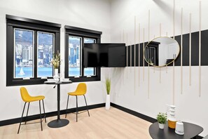 A modern hotel room with a city view, a flat-screen TV, two yellow chairs, a round table, and a decorative mirror. - Make Memories Downtown | Walk to Petco Park & Fun! (San Diego)