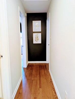 A hallway with wooden flooring, white walls, and two framed leaf designs on the wall. - Arcade Shuffleboard Poker Firepit Grill @ Battery (Marietta)