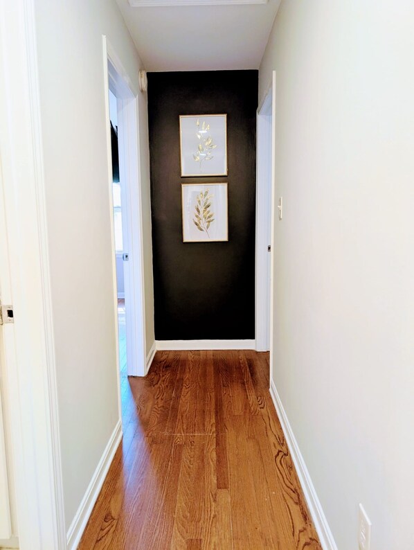 A hallway with wooden flooring, white walls, and two framed leaf designs on the wall. - Arcade Shuffleboard Poker Firepit Grill @ Battery (Marietta)
