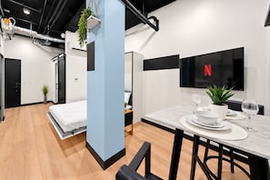 Una camera d'albergo moderna con un letto, un'area pranzo con tavolo apparecchiato per due e una TV a parete.
