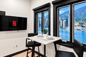 A modern room with a TV mounted on the wall, a dining table with chairs, and a view of a cityscape and mountains through large windows. - Live the Action | Walk to Petco Park Gaslamp & Fun (San Diego)