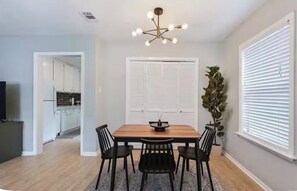 A dining area with a wooden table, four chairs, a chandelier, a potted plant, and a view of a kitchen through an open door. - Charming 2-bedroom house in beautiful College Station 2 miles from campus. (College Station)