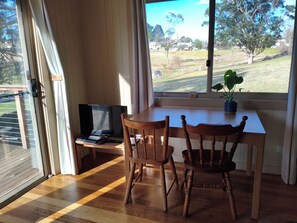 Una habitación con una mesa de madera, dos sillas, una computadora portátil y un televisor. Se ve una ventana con vistas a un área cubierta de césped y árboles.