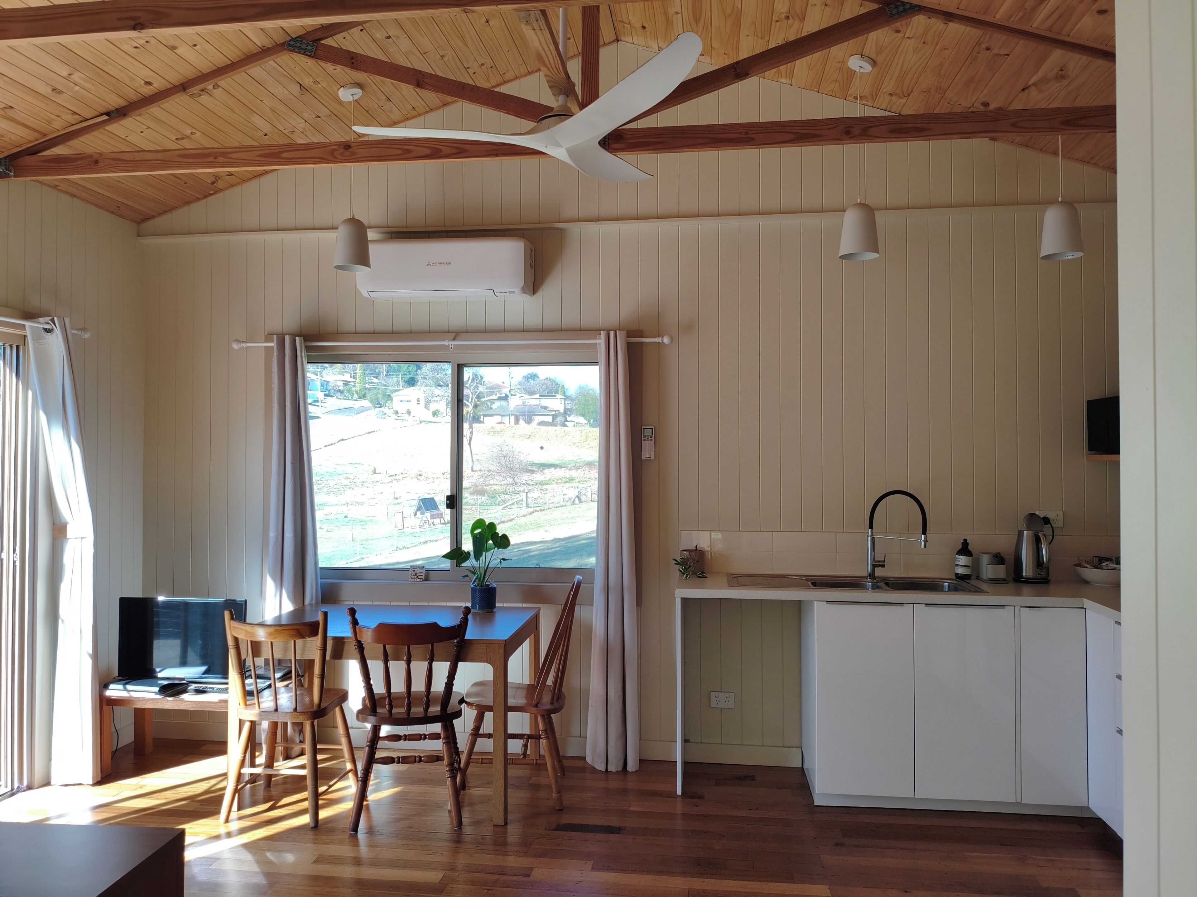 Ein Zimmer mit Esstisch, Stühlen, Deckenventilator, Klimaanlage, einem Fenster mit Vorhängen und einer Küchenzeile mit Spüle und Schränken.