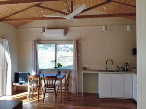 Una habitación con mesa de comedor, sillas, ventilador de techo, unidad de aire acondicionado, ventana con cortinas y una zona de cocina con lavaplatos y armarios.