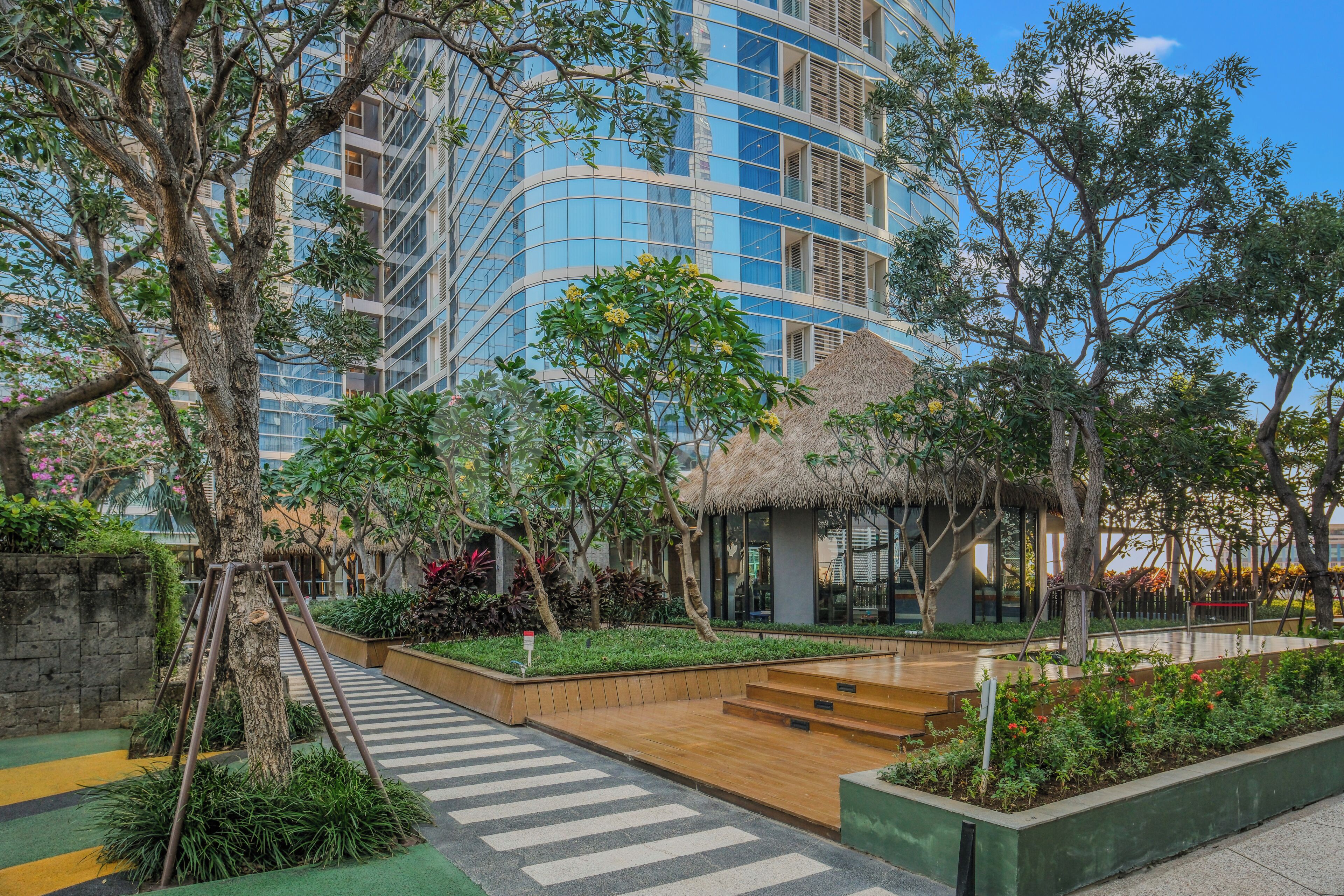 A modern hotel with a courtyard featuring a thatched-roof structure, lush greenery, and a wooden pathway.