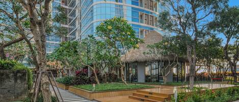 A modern hotel with a courtyard featuring a thatched-roof structure, lush greenery, and a wooden pathway.