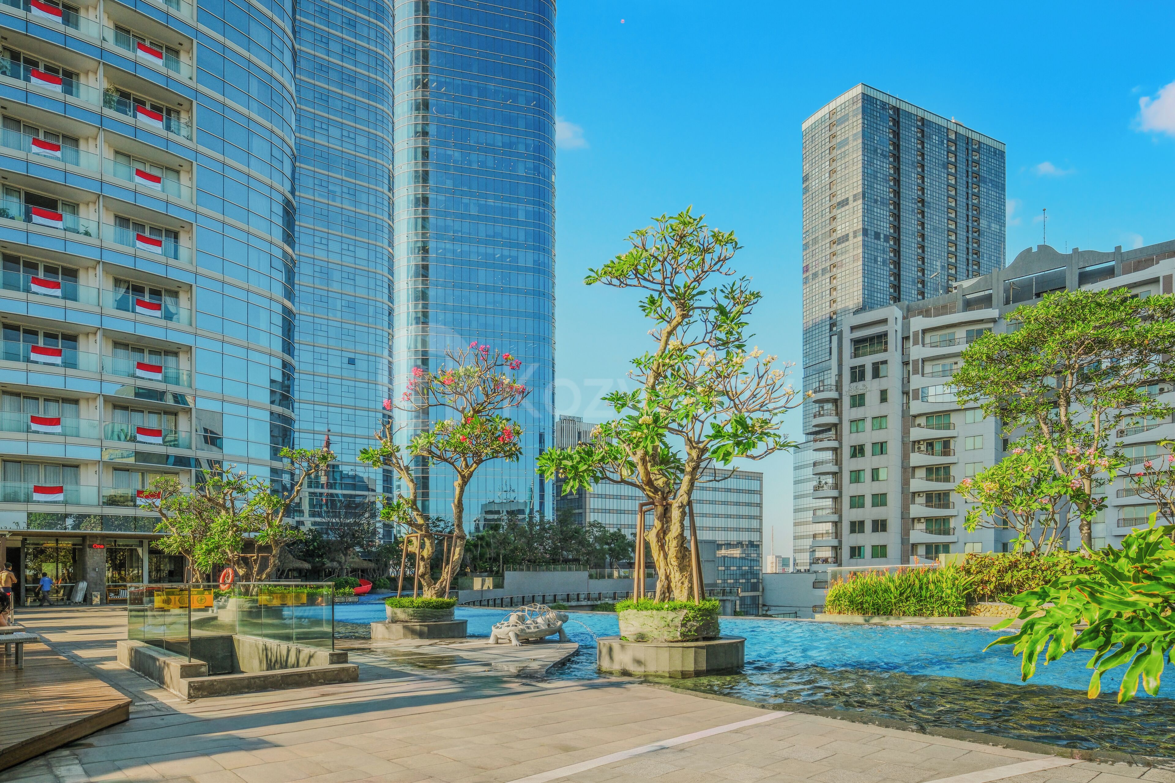 A modern urban setting with high-rise buildings, a central water feature, and greenery.