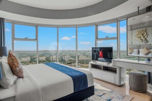 A modern bedroom with a large bed, a TV, and a desk with a view of the city. - Ardana by Kozystay - Tunjungan (Surabaya)