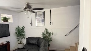 Uma sala de estar com sofá de couro preto, duas plantas em vasos, um ventilador de teto, um quadro emoldurado e uma escada.