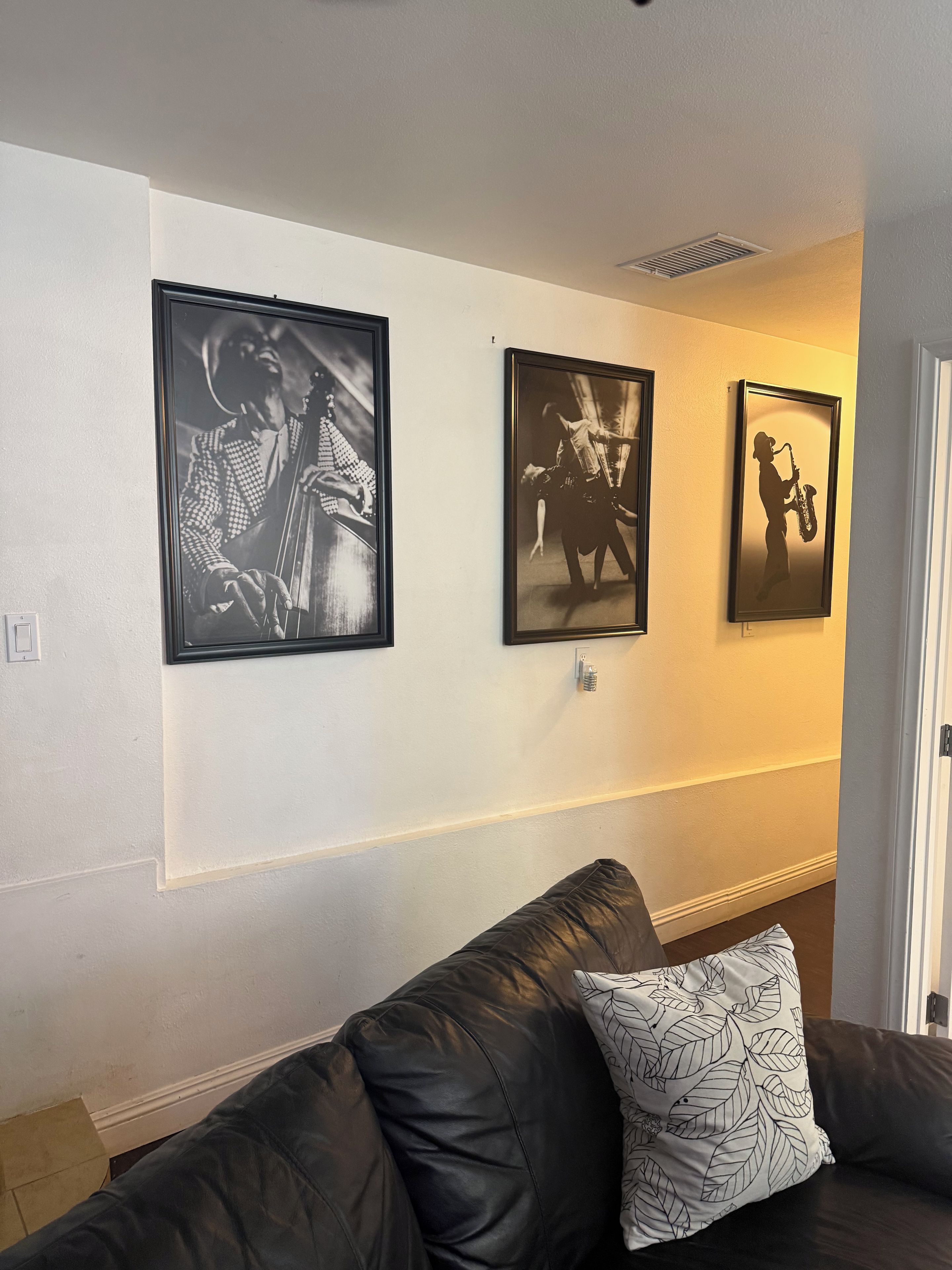 Una sala de estar con un sofá de cuero negro, un cojín estampado y tres fotografías en blanco y negro enmarcadas en la pared.