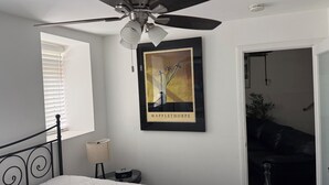 Quarto com cama, ventilador de teto, quadro emoldurado, mesa de cabeceira, abajur e janela com persianas.