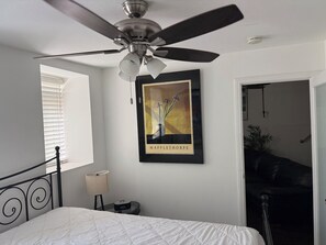 Ein Schlafzimmer mit einem Bett, einem Deckenventilator, einem gerahmten Bild, einem Nachttisch, einer Lampe und einem Fenster mit Jalousien.