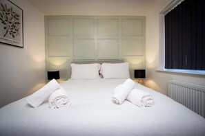 A hotel room with a large bed, two bedside lamps, a radiator, and a window with blinds. - Stylish 2BR Flat in Tyldesley with Parking & Sleeps 4 (Tyldesley)