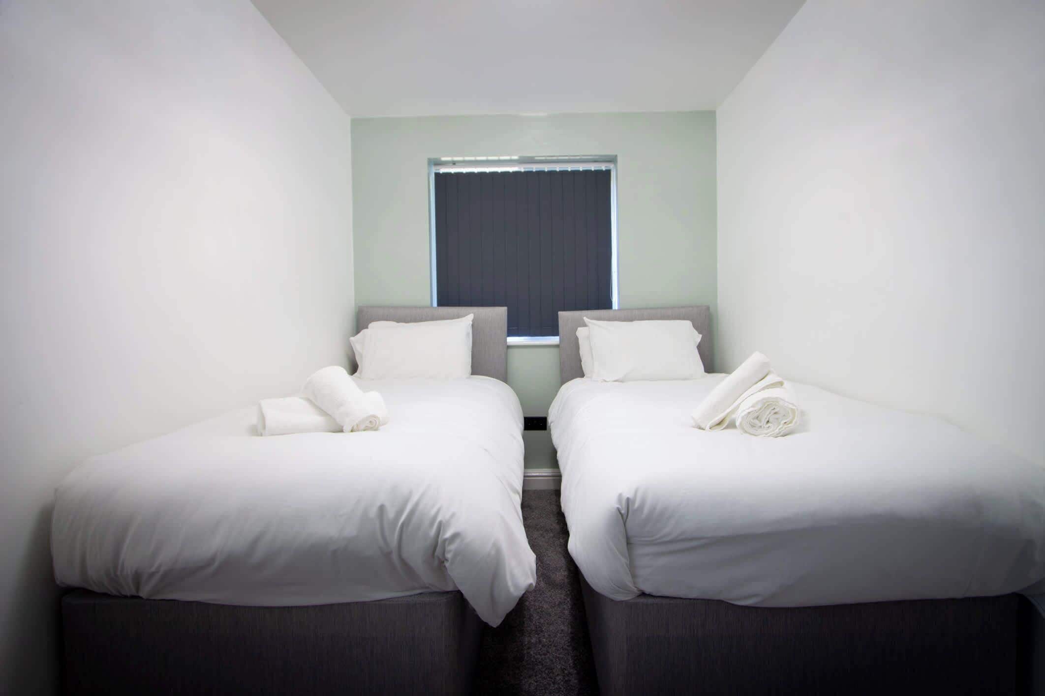 A hotel room with two single beds, a window with blinds, and a carpeted floor.