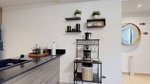 Una cocina moderna con encimera negra, cafetera y pileta. Se observan estantes con plantas y utensilios de cocina.