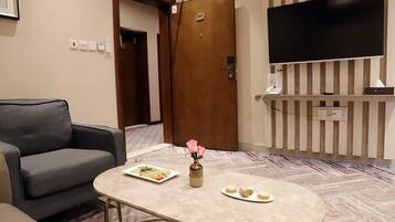 A hotel room with a sofa, a table with a vase and a plate of food, a TV mounted on the wall, and a door with a number plate.