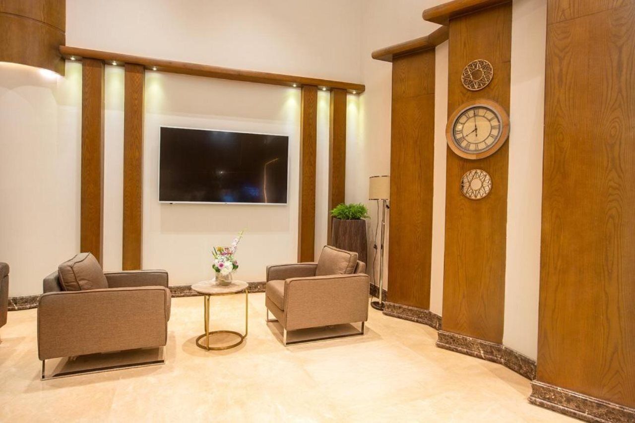 Um lobby moderno com televisão de tela plana, dois sofás, uma mesa redonda pequena com um vaso e um pilar de madeira com relógios.