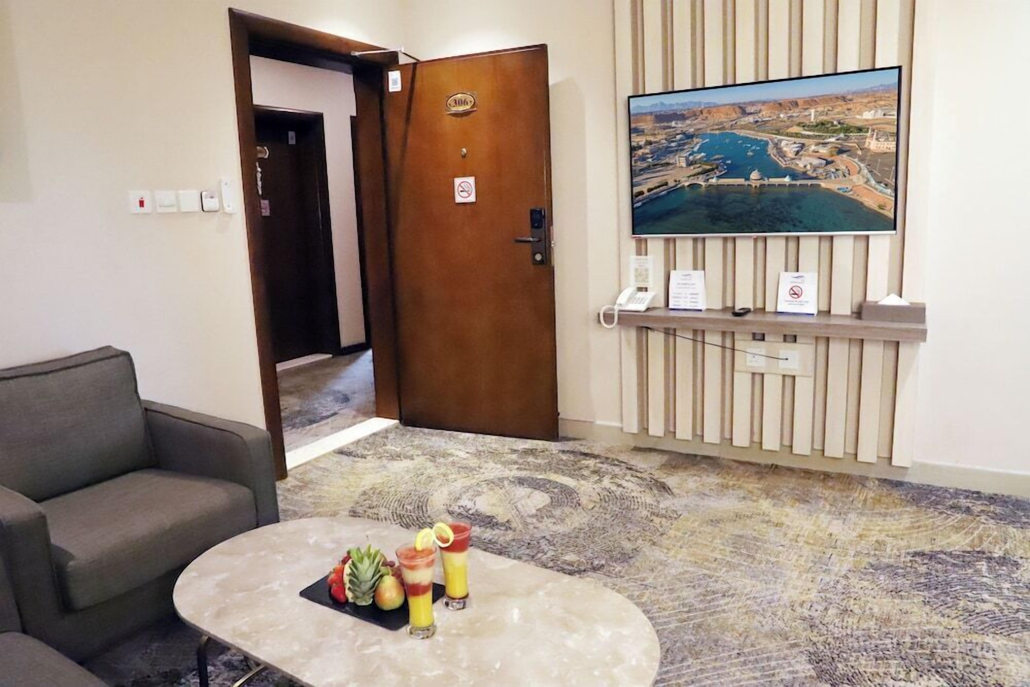 Um hall de hotel com uma mesa de recepção, uma televisão exibindo uma vista urbana, um sofá e uma mesa de centro com frutas e bebidas.