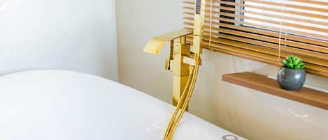 A bathtub with rose petals, a gold faucet, and a small plant on a shelf.