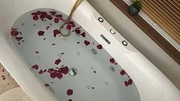 Une baignoire dans laquelle flottent des pétales de rose, un robinet doré et une petite plante en pot posée sur le rebord de la fenêtre.