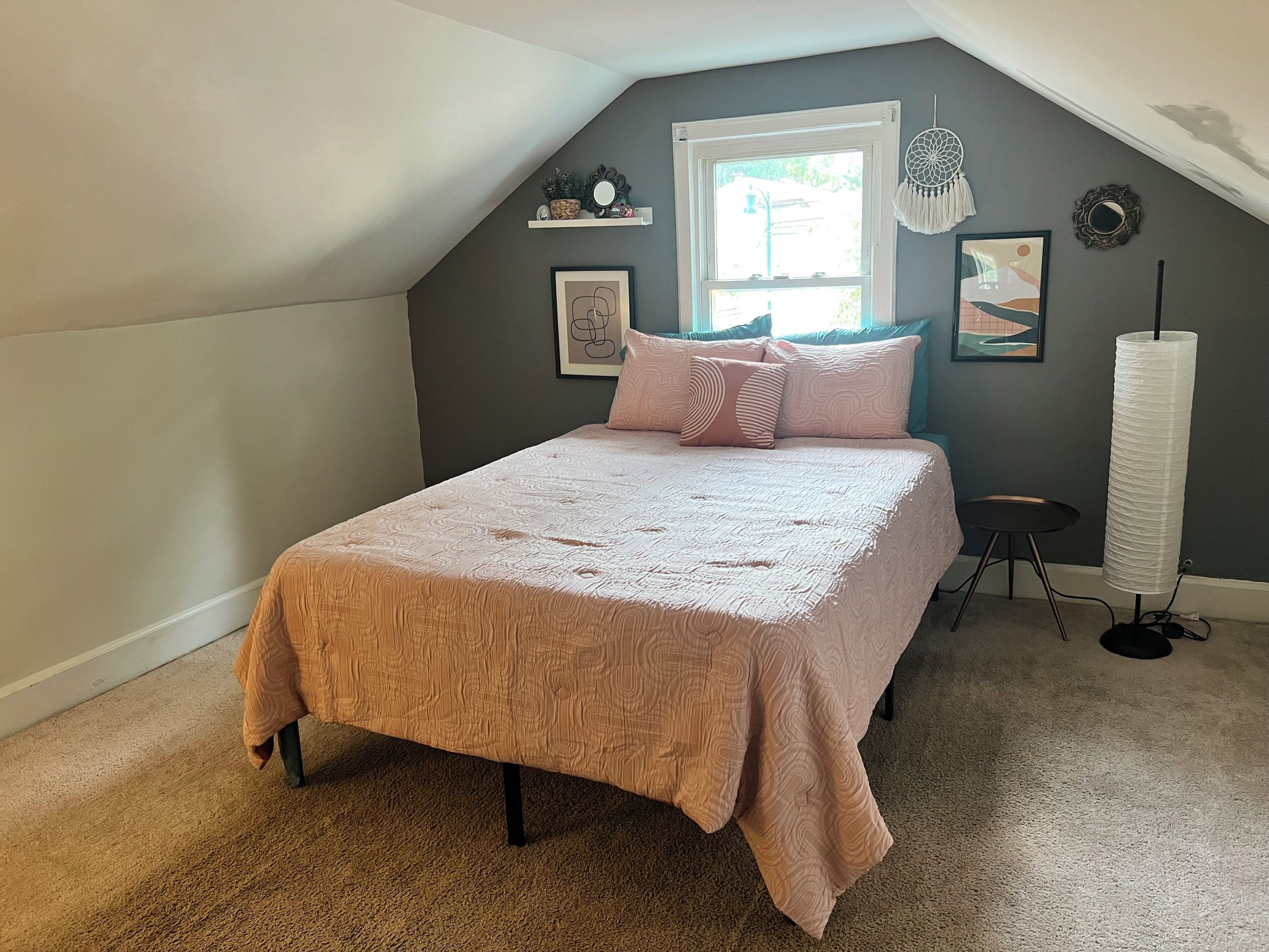 A bedroom with a sloped ceiling, a bed with a patterned bedspread, a window with a view, and a bedside table with a lamp.