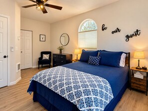 Une chambre avec un lit, deux tables de chevet, une chaise, un ventilateur de plafond, une fenêtre avec des stores et un panneau décoratif mural.