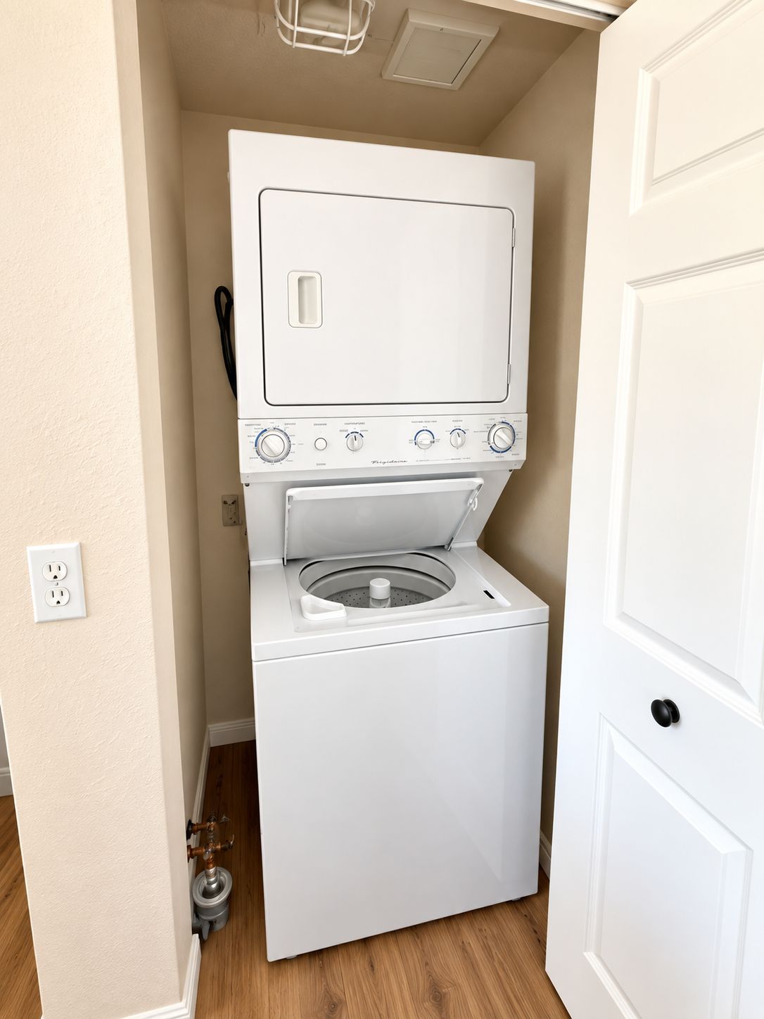Un appareil compact, blanc, combinant une machine à laver et un sèche-linge à chargement frontal, installé dans une petite buanderie avec un plancher en bois et une porte blanche.