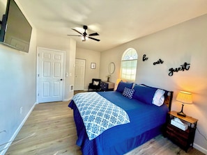 A bedroom with a bed, a nightstand, a ceiling fan, a window with a view, and a wall sign.