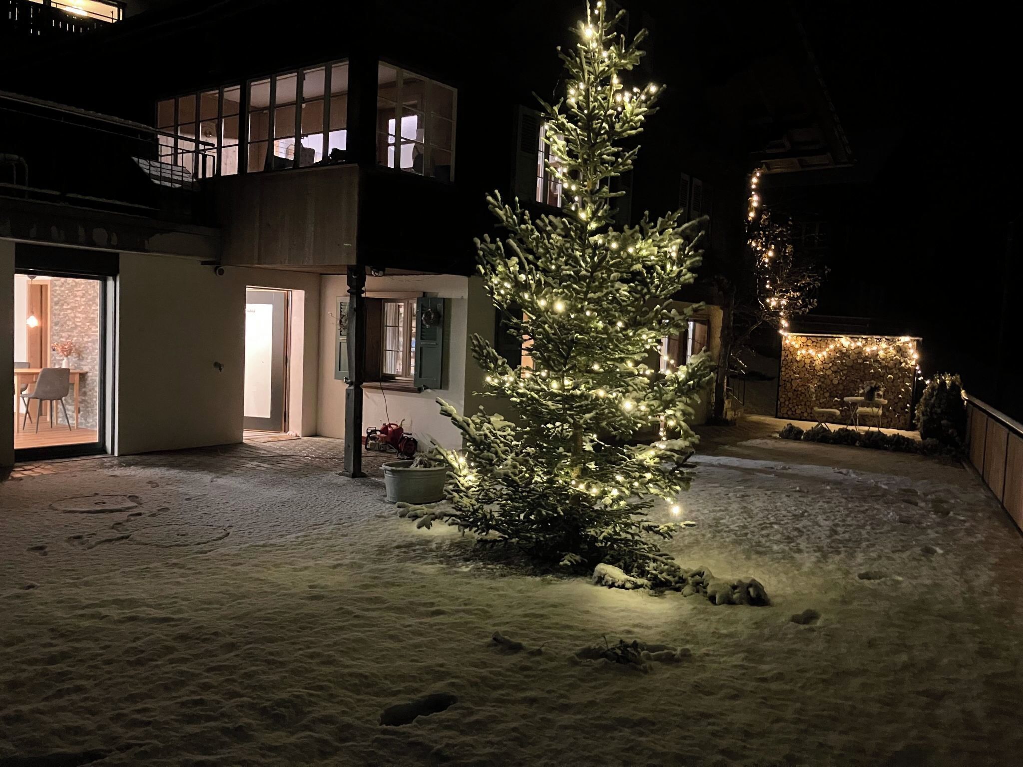 En snedækket gårdsplads med et oplyst juletræ, en bygning med mange vinduer og en mur med lys.