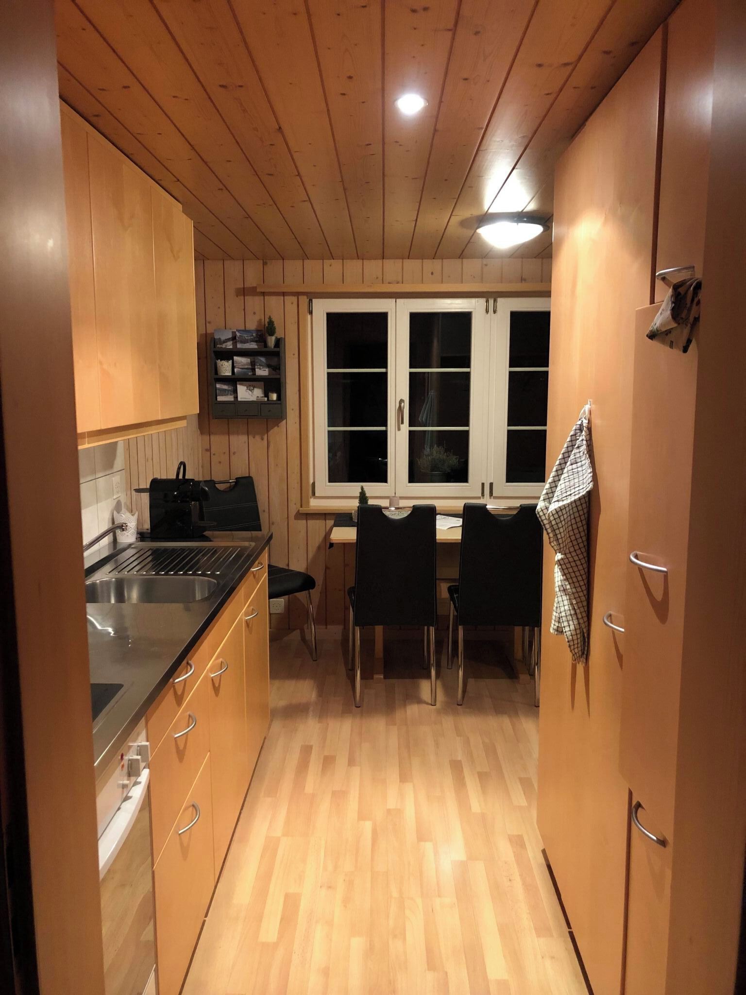 Een kleine keuken met houten kastjes, een zwart aanrecht en een eethoek met twee stoelen.