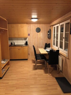 Eine kleine Küche mit Holzschränken, einem Esstisch mit Stühlen und einem Fenster mit einer Uhr.