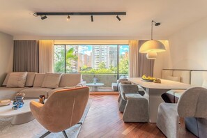 Ein modernes Wohnzimmer mit einer Couch, Sesseln, einem Esstisch und Blick auf Gebäude durch große Fenster.