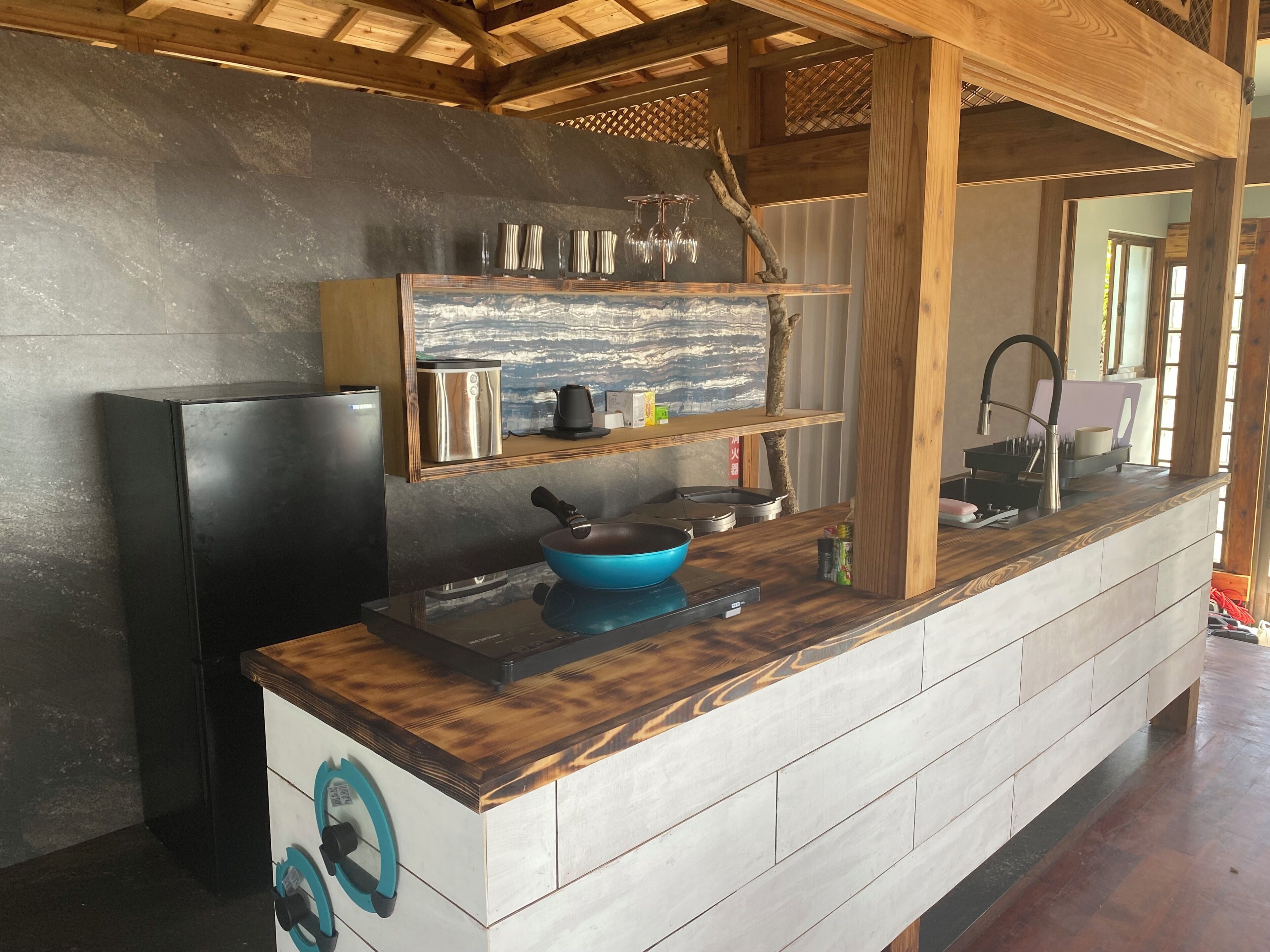 Una cucina con un frigorifero nero, una pentola da cucina blu, una macchina per il caffè e una mensola in legno con bicchieri.
