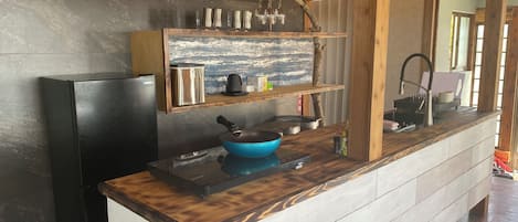 Une cuisine avec un réfrigérateur noir, une casserole bleue, une machine à café et une étagère en bois où sont rangés des verres.