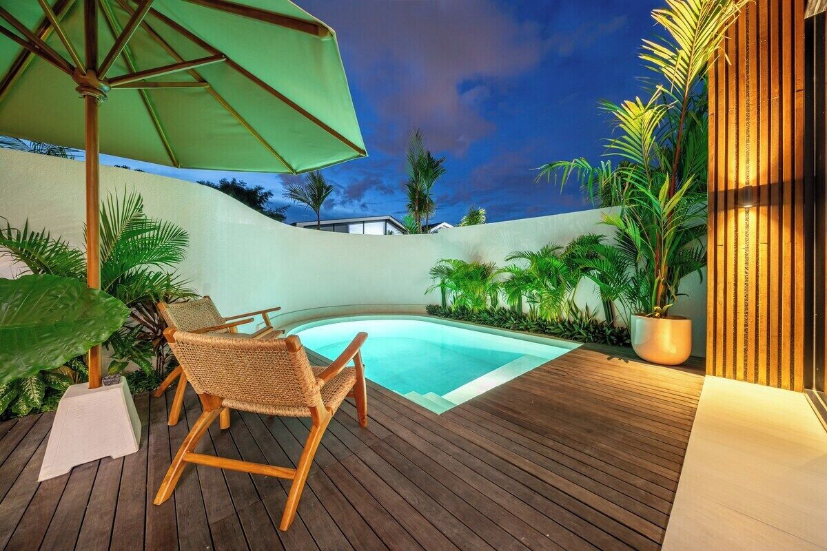 Ein Holzdeck mit einem Pool, umgeben von tropischen Pflanzen und einem grünen Sonnenschirm.