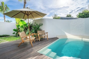 Ein Schwimmbecken mit Holzterrasse und Korbstühlen, umgeben von üppiger Vegetation.