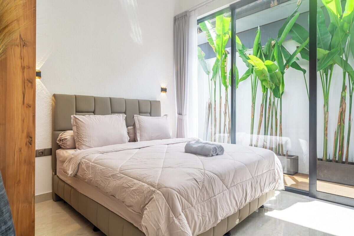 Une chambre moderne avec un grand lit, une tête de lit et une vue sur des plantes tropicales, visible à travers une porte vitrée.