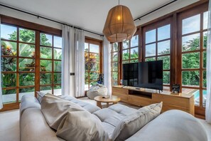 A modern living room with a sofa, TV, and large windows overlooking greenery. - Hidden 3BR Oasis in Serene Ubud w Huge Pool+Patio (Gianyar Regency)