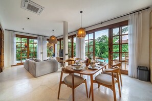 A spacious living area with a dining table, chairs, and a sofa. Large windows provide a view of greenery outside. - Hidden 3BR Oasis in Serene Ubud w Huge Pool+Patio (Gianyar Regency)