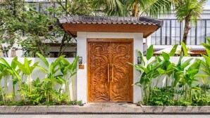 A traditional wooden gate with intricate carvings, flanked by lush green plants and a white wall. - Cozy 3BR in Ubud w Bathtubs, Ricefield Views, Pool (Tegallalang)
