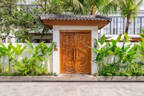 Um portão de madeira tradicional com entalhes elaborados, ladeado por plantas verdes exuberantes e uma parede branca.