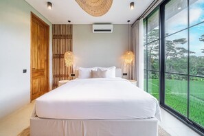Ein Schlafzimmer mit einem großen Bett, zwei Nachttischlampen, einer Deckenleuchte und Blick ins Grüne durch ein großes Fenster.