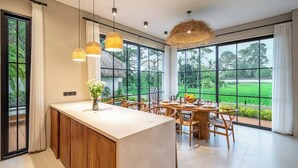 A modern kitchen with a large island, dining area, and large windows with a view of greenery. - Luxe 3BR Gem in Tropical Ubud w Pool, Patio&Garden (Gianyar Regency)